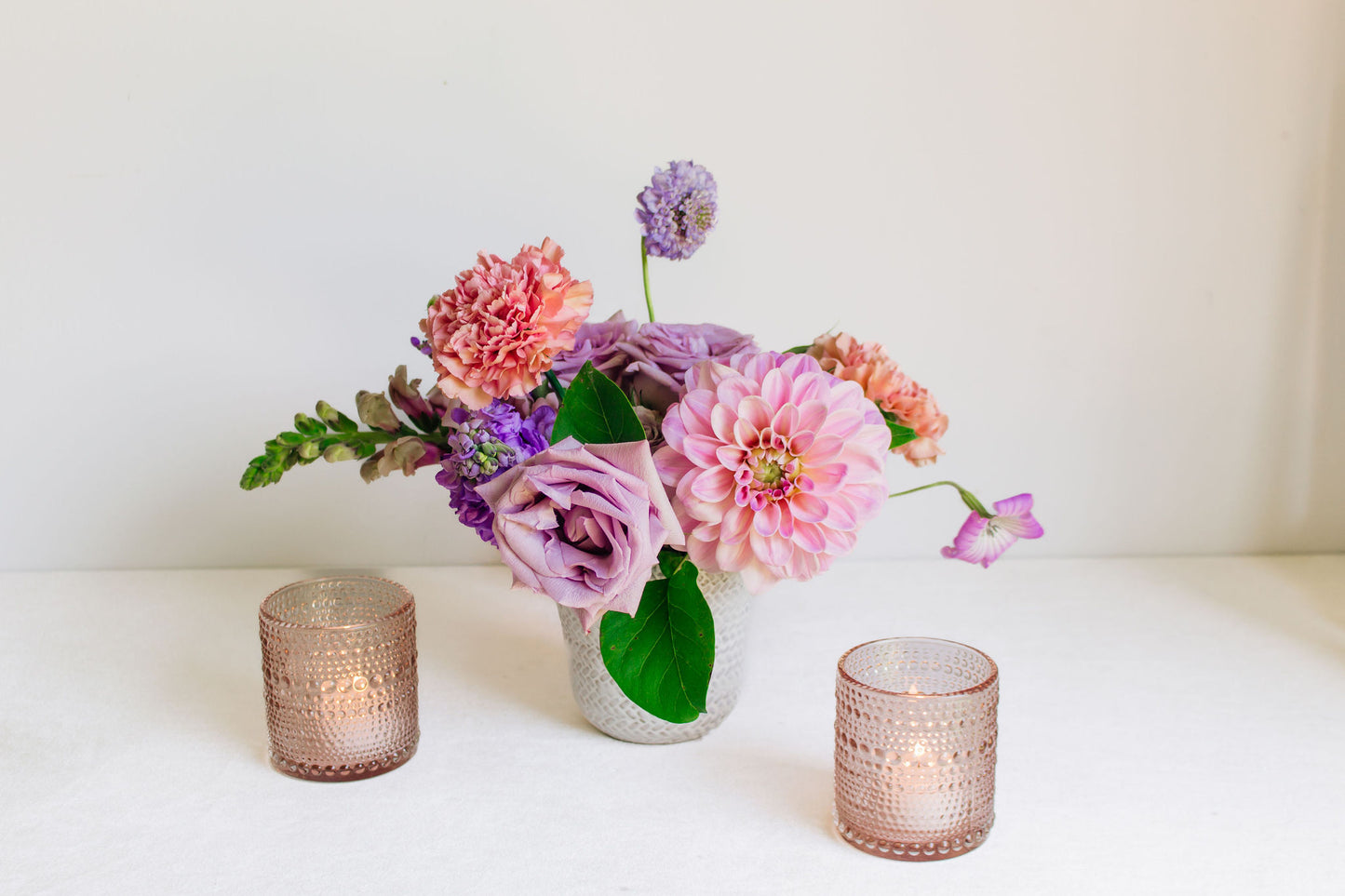 Petite white vase filled with light purple and pink flowers, such as dahlias, roses, carnations, and more. Paired with blush bubble votives. 