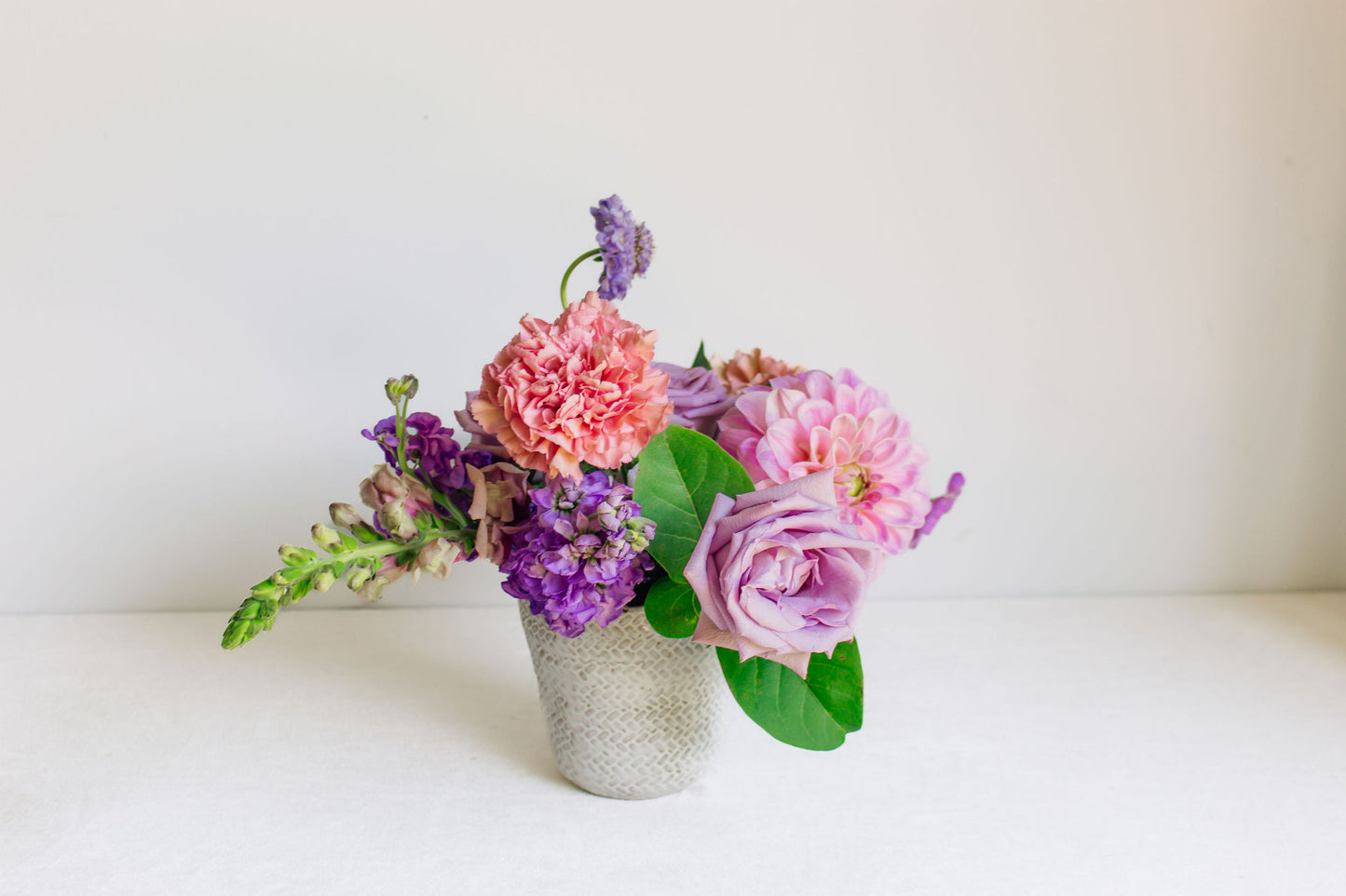 Petite white vase filled with light purple and pink flowers, such as dahlias, roses, carnations, and more. 