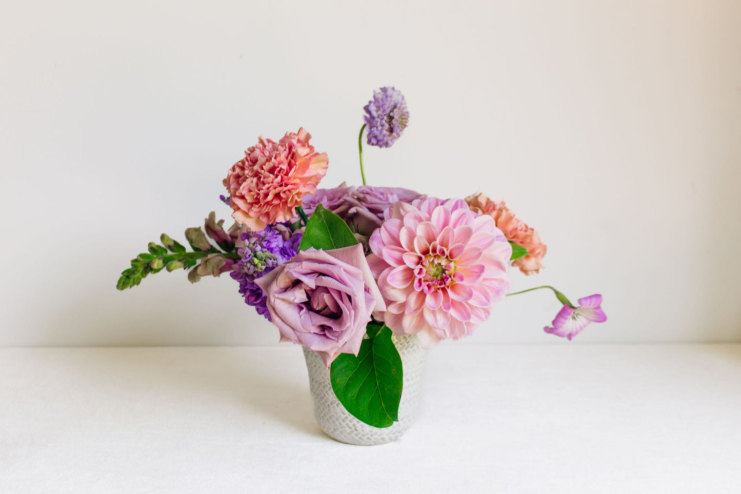 Petite white vase filled with light purple and pink flowers, such as dahlias, roses, carnations, and more.