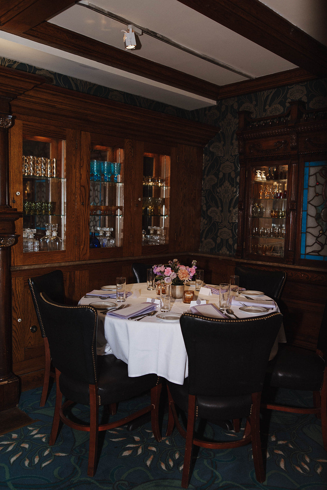 Dining table set for four with purple and pink floral centerpieces in a dimly lit room with wooden beams and shelves.