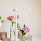 Trio of clear glass bud vases of light purple and light pink flowers, such as dahlias, stock, carnations, roses, scabiosa, and corncockle. White taper candles and light pink votives interspersed throughout. 
