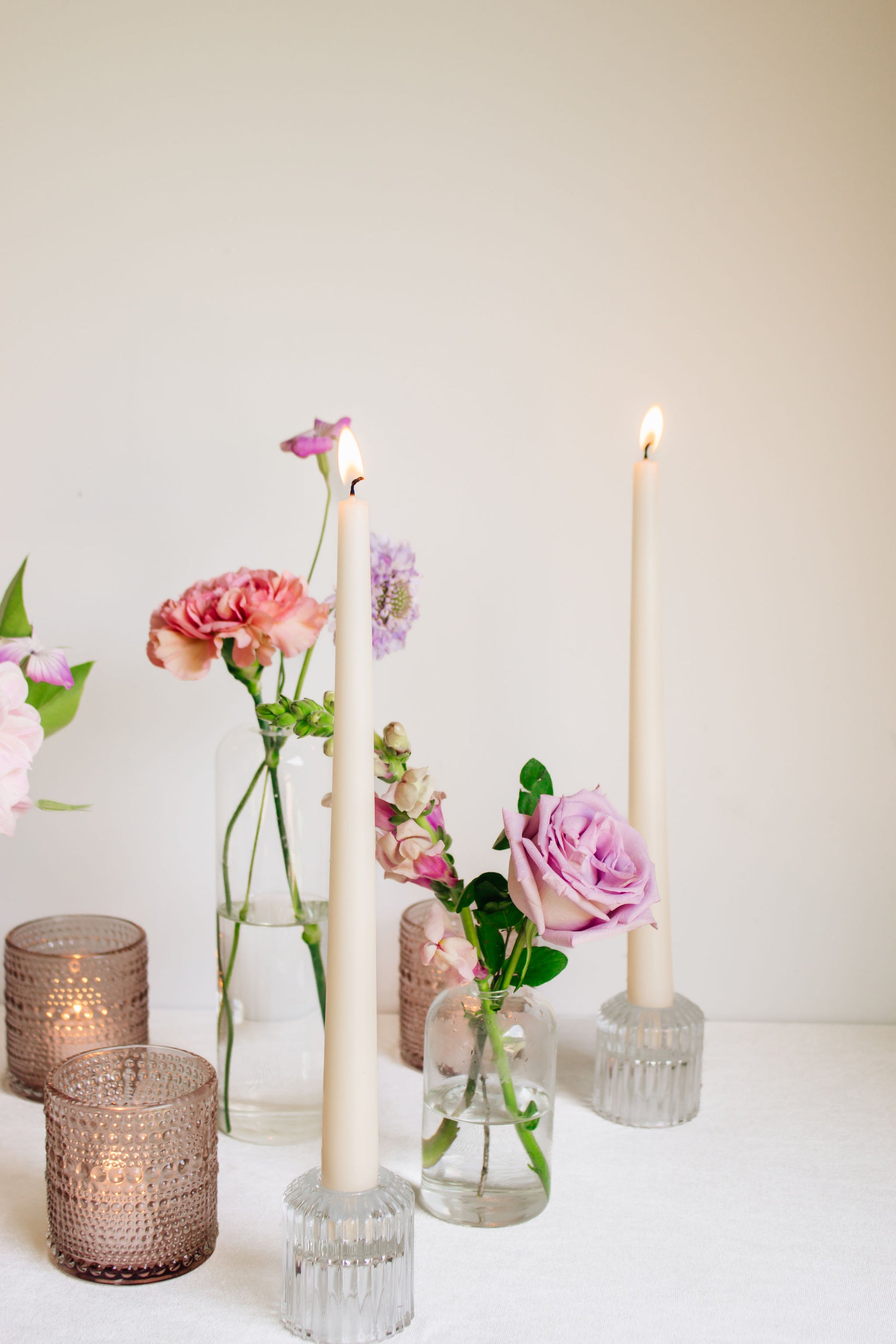 Trio of clear glass bud vases of light purple and light pink flowers, such as dahlias, stock, carnations, roses, scabiosa, and corncockle. White taper candles and light pink votives interspersed throughout. 