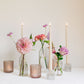 Trio of clear glass bud vases of light purple and light pink flowers, such as dahlias, stock, carnations, roses, scabiosa, and corncockle. White taper candles and light pink votives interspersed throughout. 