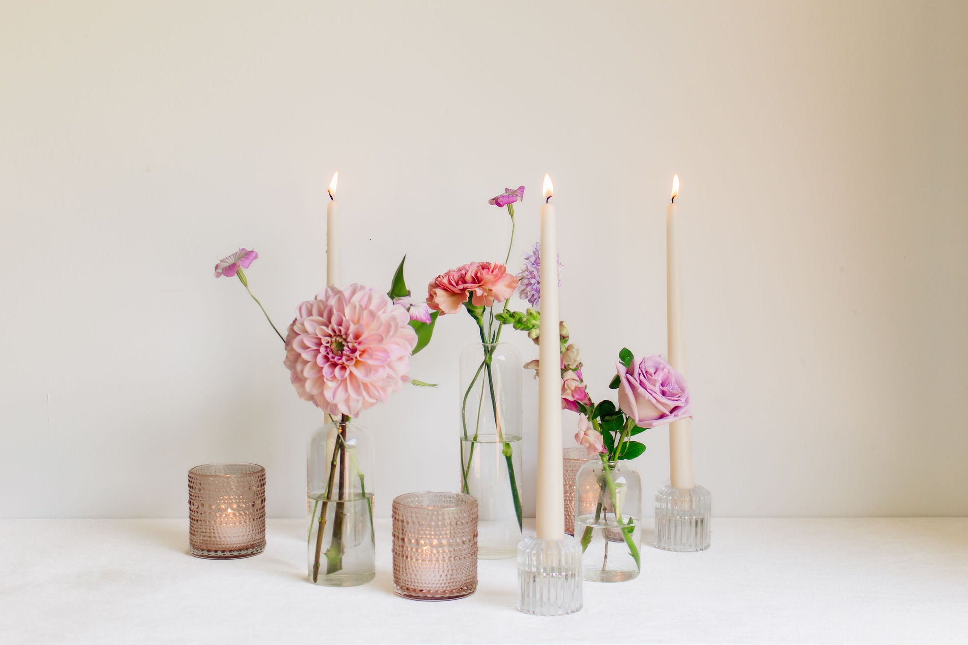 Trio of clear glass bud vases of light purple and light pink flowers, such as dahlias, stock, carnations, roses, scabiosa, and corncockle. White taper candles and light pink votives interspersed throughout. 