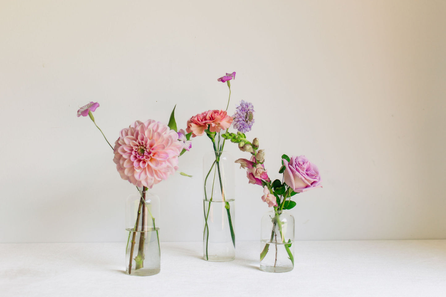 Trio of clear glass bud vases of light purple and light pink flowers, such as dahlias, stock, carnations, roses, scabiosa, and corncockle. 