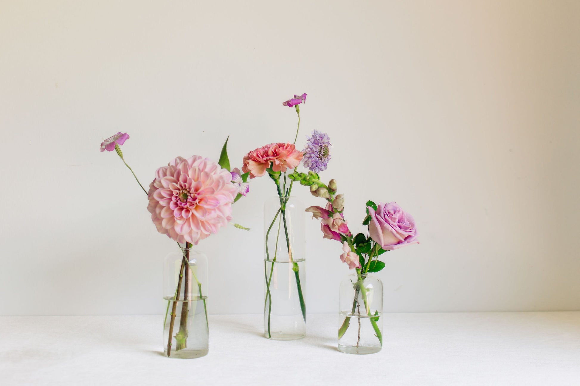 Trio of clear glass bud vases of light purple and light pink flowers, such as dahlias, stock, carnations, roses, scabiosa, and corncockle. 