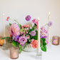 Centerpiece in a white bowl of a garden style arrangement of light purple and light pink flowers, such as dahlias, stock, carnations, roses, scabiosa, and corncockle. White taper candles and light pink votives surround the arrangement. 