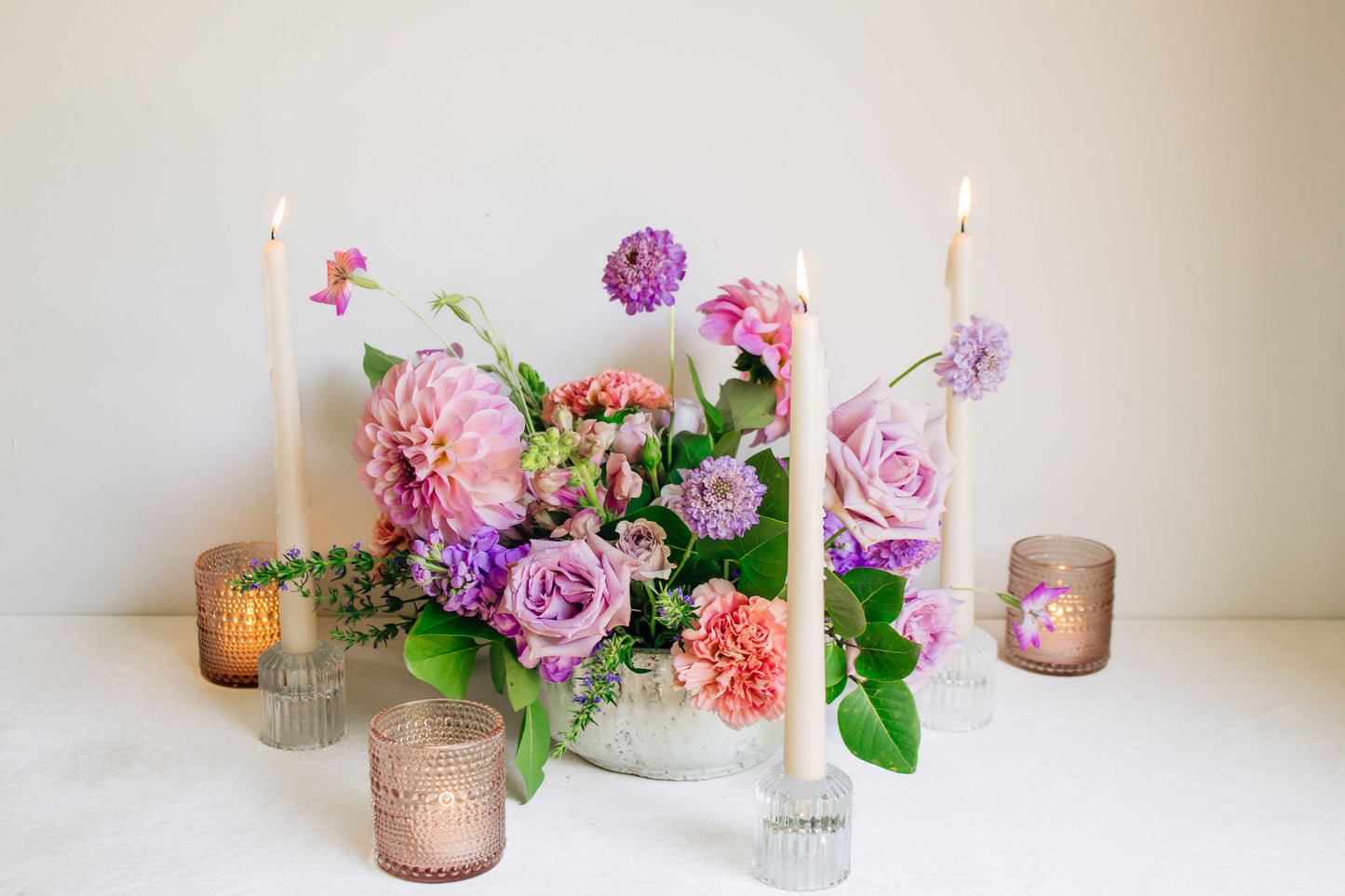 Centerpiece in a white bowl of a garden style arrangement of light purple and light pink flowers, such as dahlias, stock, carnations, roses, scabiosa, and corncockle. White taper candles and light pink votives surround the arrangement. 