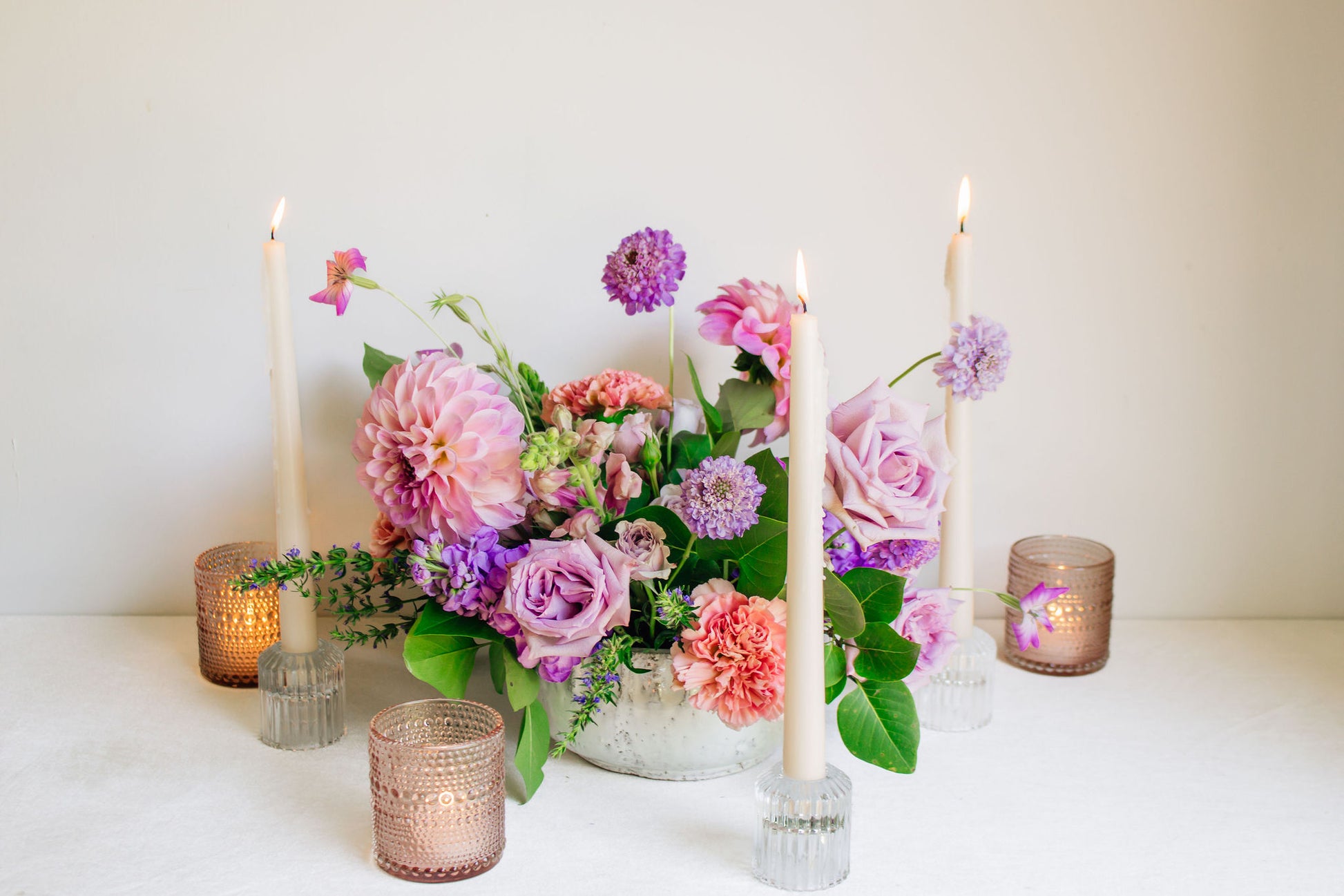 Centerpiece in a white bowl of a garden style arrangement of light purple and light pink flowers, such as dahlias, stock, carnations, roses, scabiosa, and corncockle. White taper candles and light pink votives surround the arrangement. 