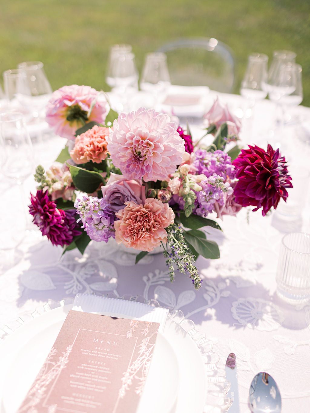 Centerpiece in a white bowl of a garden style arrangement of purple and light pink flowers, such as dahlias, stock, carnations, mums, and spray roses. 