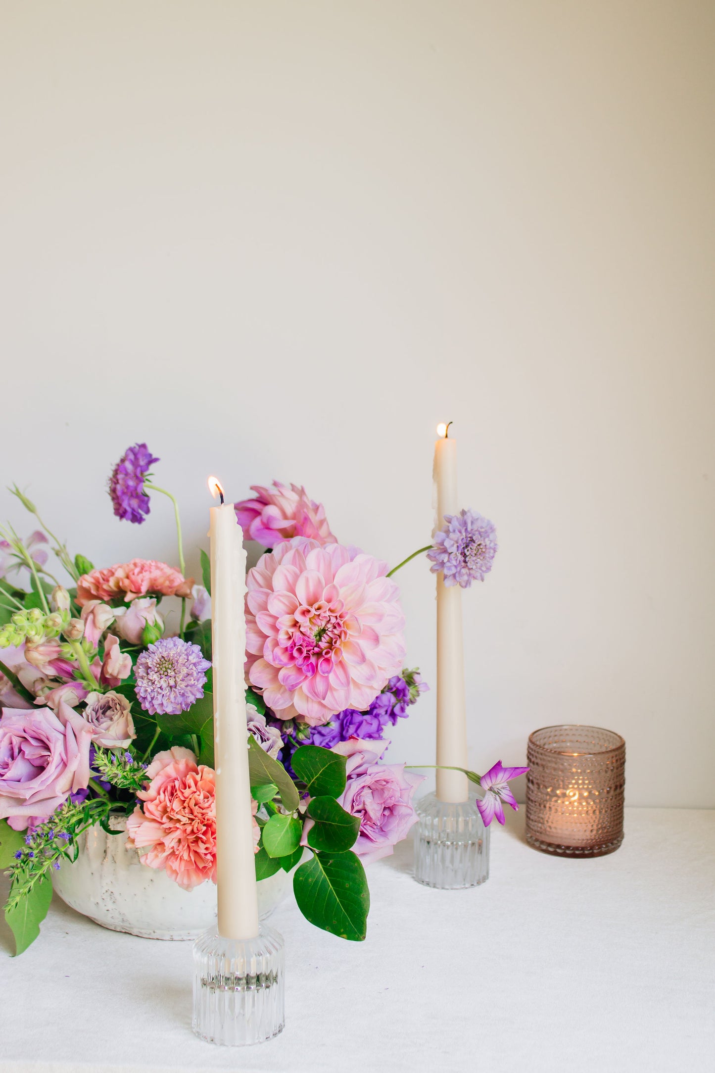 Centerpiece in a white bowl of a garden style arrangement of light purple and light pink flowers, such as dahlias, stock, carnations, roses, scabiosa, and corncockle. White taper candles and light pink votives surround the arrangement. 
