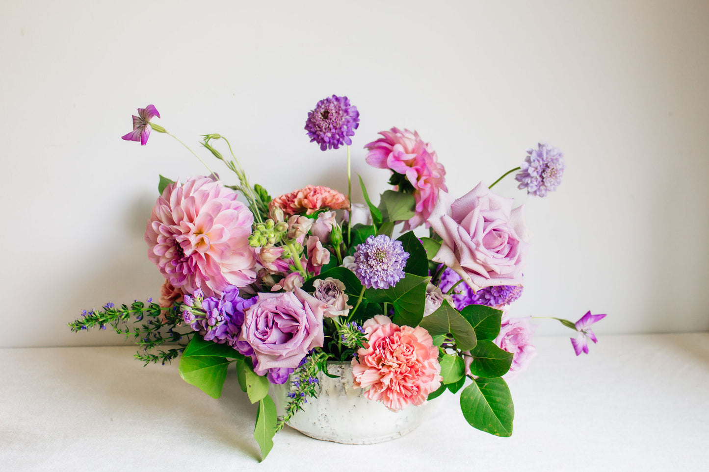 Centerpiece in a white bowl of a garden style arrangement of light purple and light pink flowers, such as dahlias, stock, carnations, roses, scabiosa, and corncockle. 