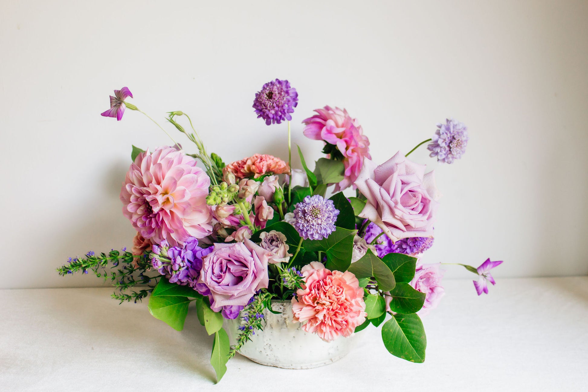 Centerpiece in a white bowl of a garden style arrangement of light purple and light pink flowers, such as dahlias, stock, carnations, roses, scabiosa, and corncockle. 