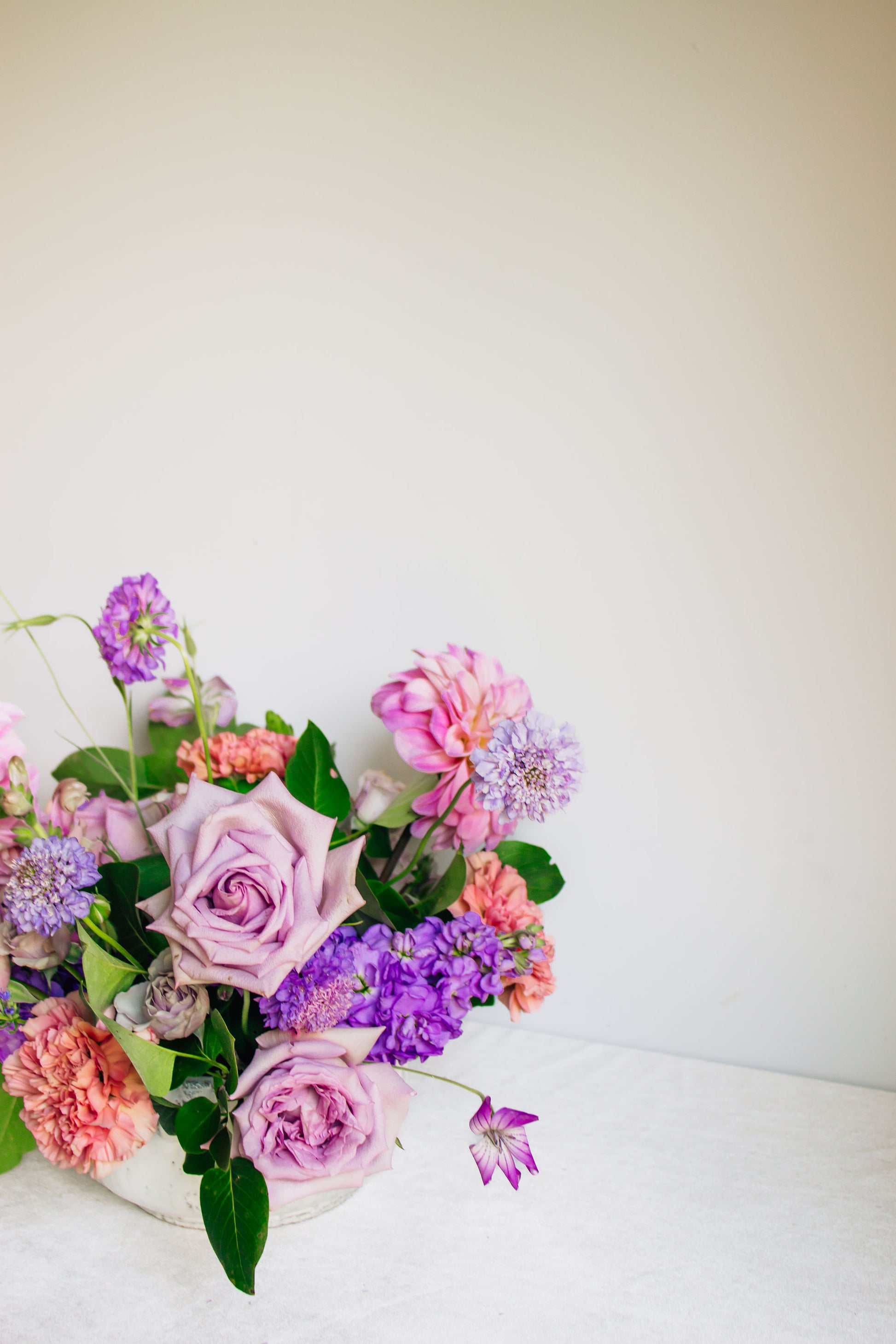 Centerpiece in a white bowl of a garden style arrangement of light purple and light pink flowers, such as dahlias, stock, carnations, roses, scabiosa, and corncockle.