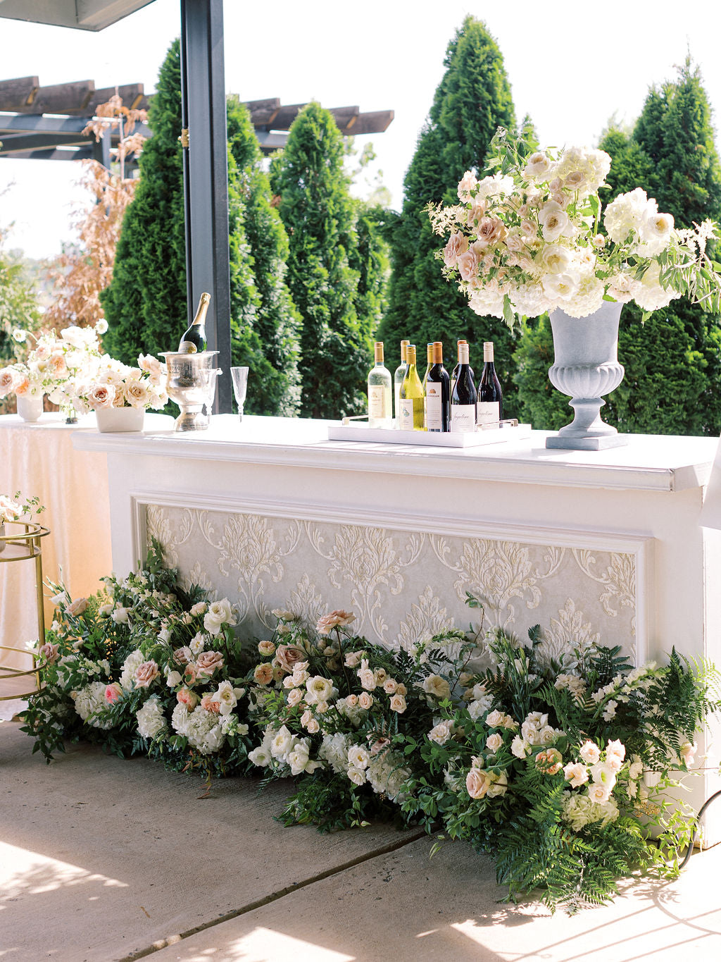 Bar at a wedding with bottles of wine and a large floral arrangement in a stone urn on the bar top. Floral arrangements on the ground line the front of the bar. Floral arrangements are made of white and blush flowers and greenery. Primarily roses in a classic garden style. The bar is white with a classic, vintage decorative front. 