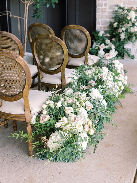 Floral arrangements on the ground lining the aisle of a wedding ceremony. Floral arrangements are primarily white, blush, and green. Ceremony chairs are cane back chairs with white cushioned seats.