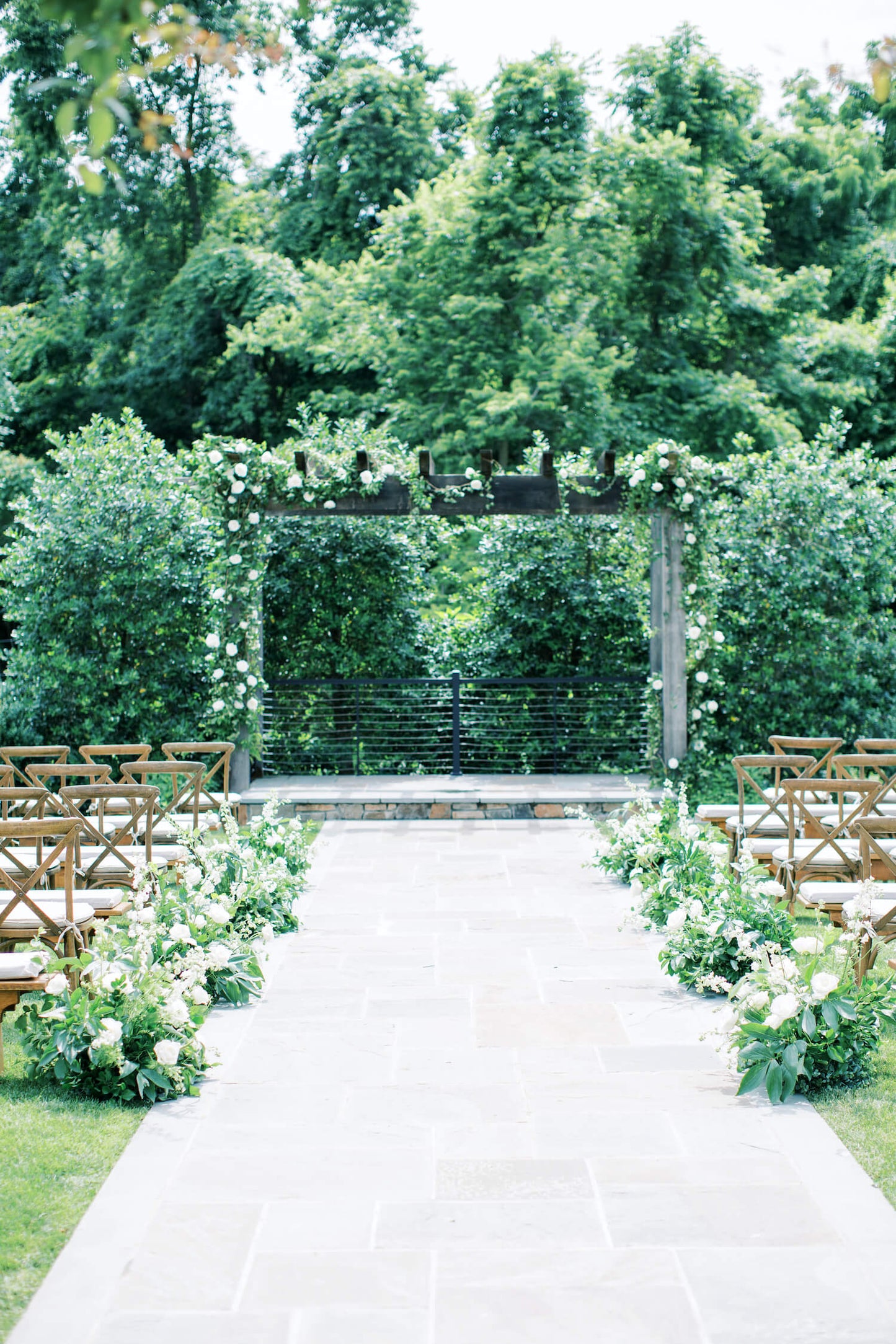 Wedding ceremony with a wooden arch with organic vining of greenery and white flowers. Floral arrangements of greenery and white flowers line the aisle.