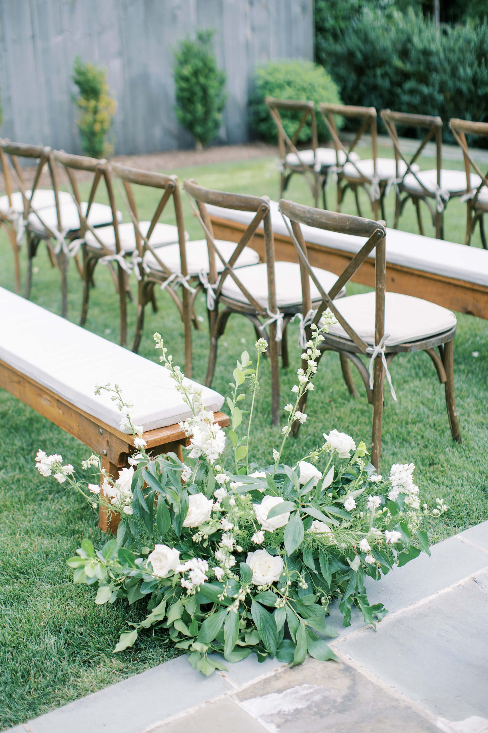 Floral arrangement of greenery and white flowers on the ground of a wedding ceremony aisle. It's a close up of the arrangement next to a wooden bench with padded seating. 