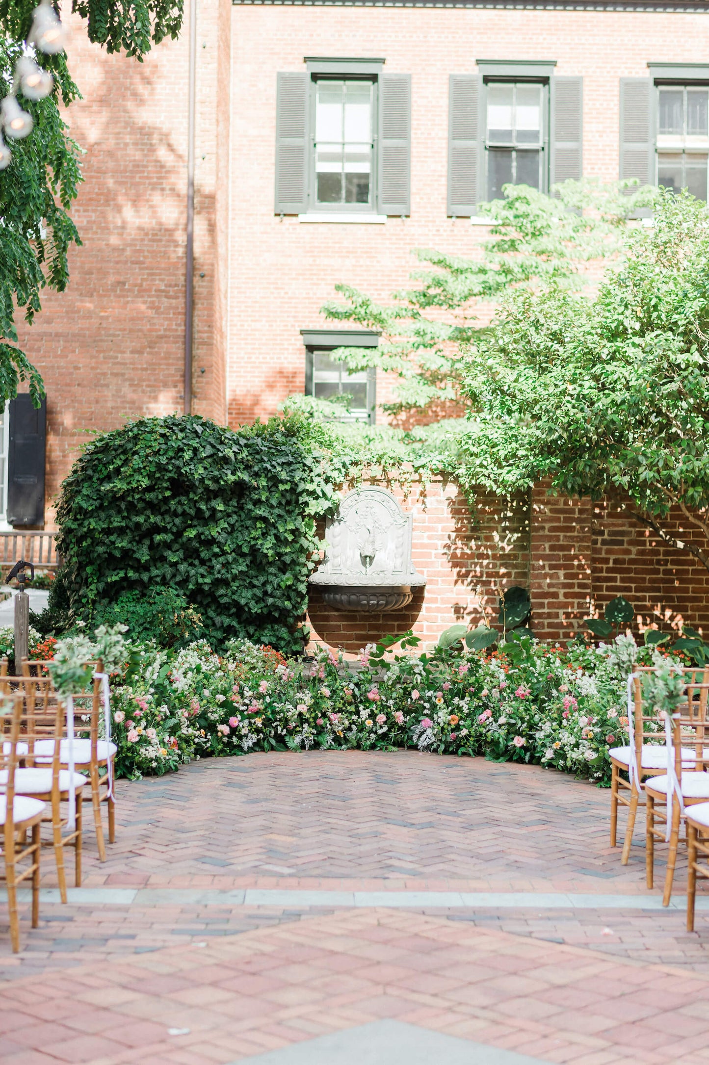 Wedding ceremony in a brick courtyard. The back brick wall has ivy growing on it a stone fountain fixed to the center of the wall. Floral arrangements on the ground form a semi-circle. The arrangements are primarily greenery with bright colored flowers, like pink and orange. Wood chiavari chairs with white seats are in the foreground. 