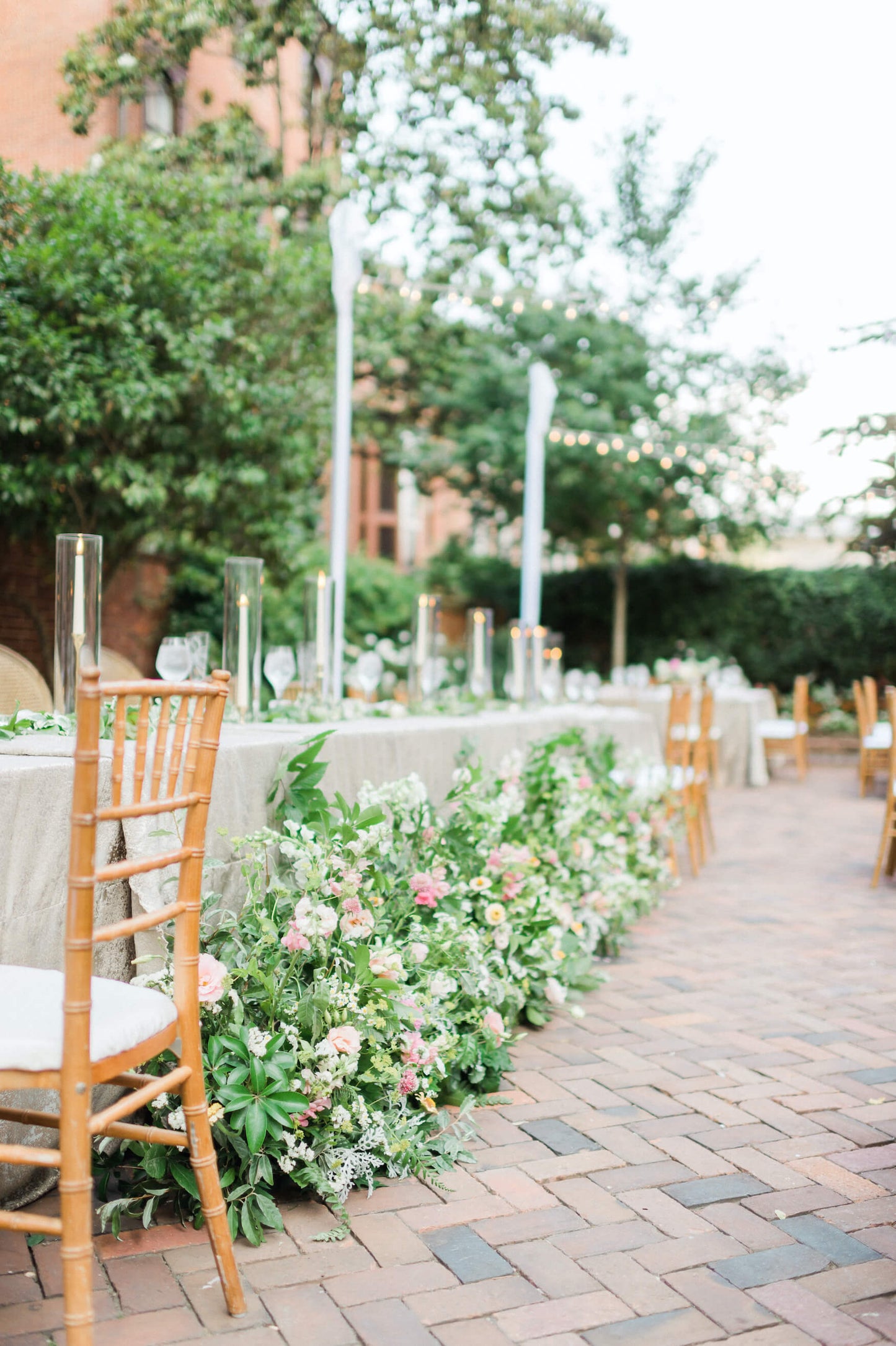 Floral arrangements on the ground line the front of. along rectangular table. The arrangements are primarily greenery with white, pink, orange, and yellow flowers. The table has a light sage linen on it and a few white taper candles. A wood chiavari chair is in the foreground. 