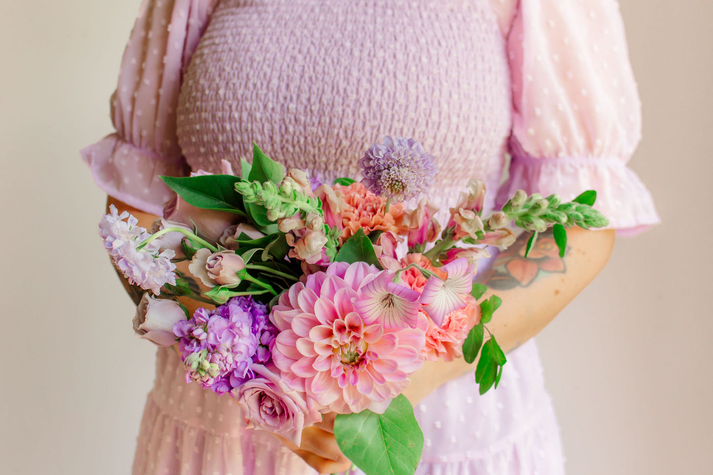 A small bouquet of lavender, mauve, and purple flowers, such as dahlias, sotck, carnations, snapdragons, and corncockle, held in a woman's hand wearing a lavender dress. 