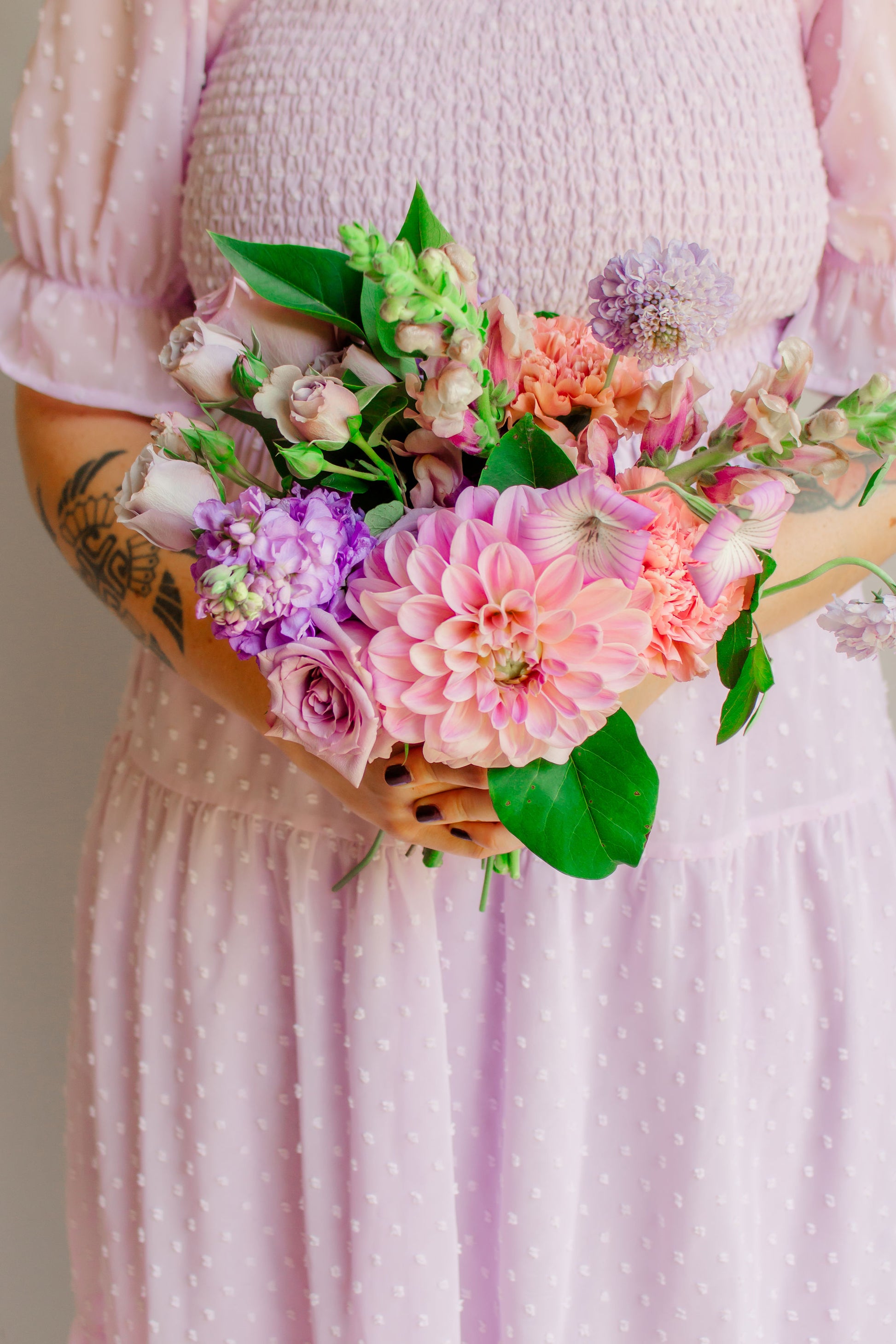 A small bouquet of lavender, mauve, and purple flowers, such as dahlias, sotck, carnations, snapdragons, and corncockle, held in a woman's hand wearing a lavender dress. 
