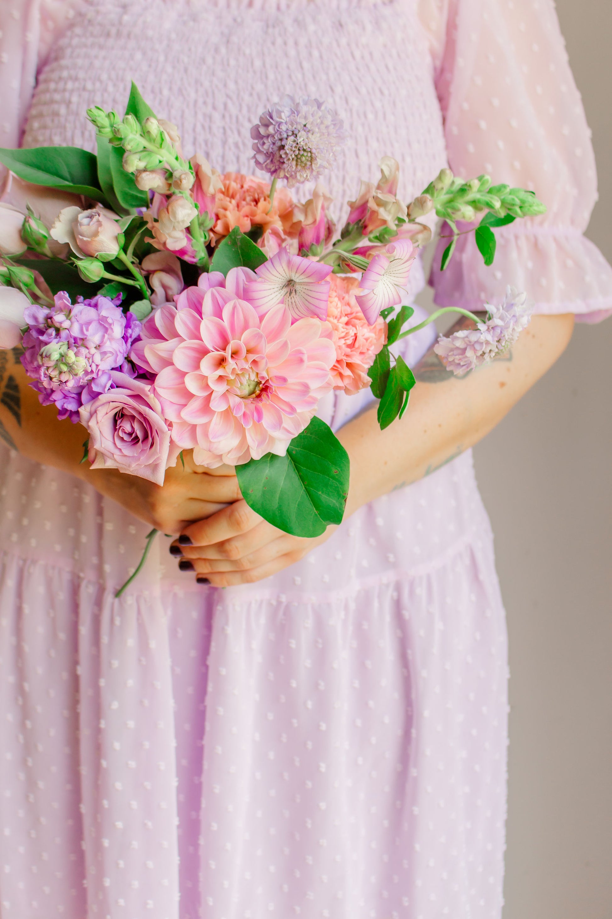 A small bouquet of lavender, mauve, and purple flowers, such as dahlias, sotck, carnations, snapdragons, and corncockle, held in a woman's hand wearing a lavender dress. 