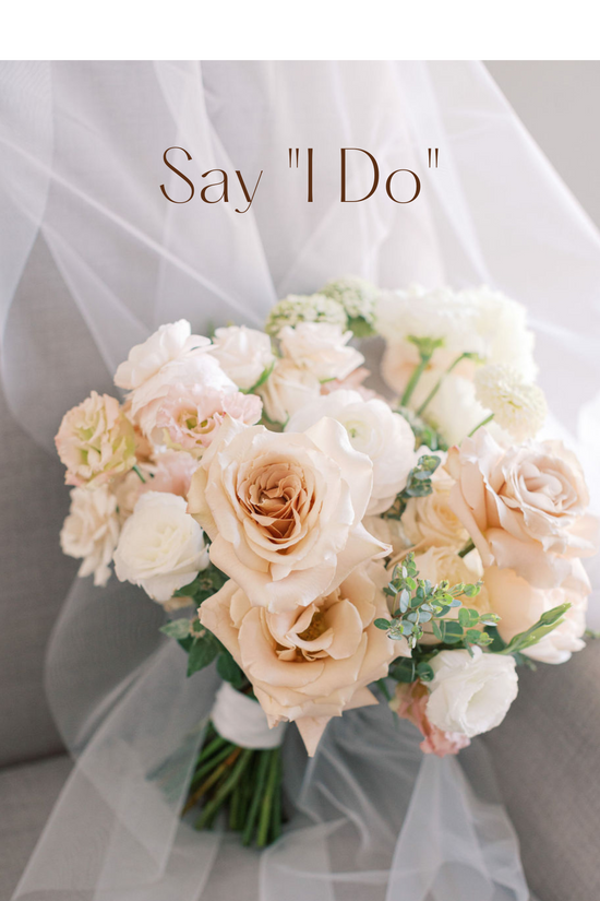 Soft blush and white bridal bouquet resting on a white veil. Bouquet is primarily made of roses, ranunculus, and lisianthus. The image says 