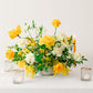 Floral arrangement with yellow and white flowers and candles on a white surface