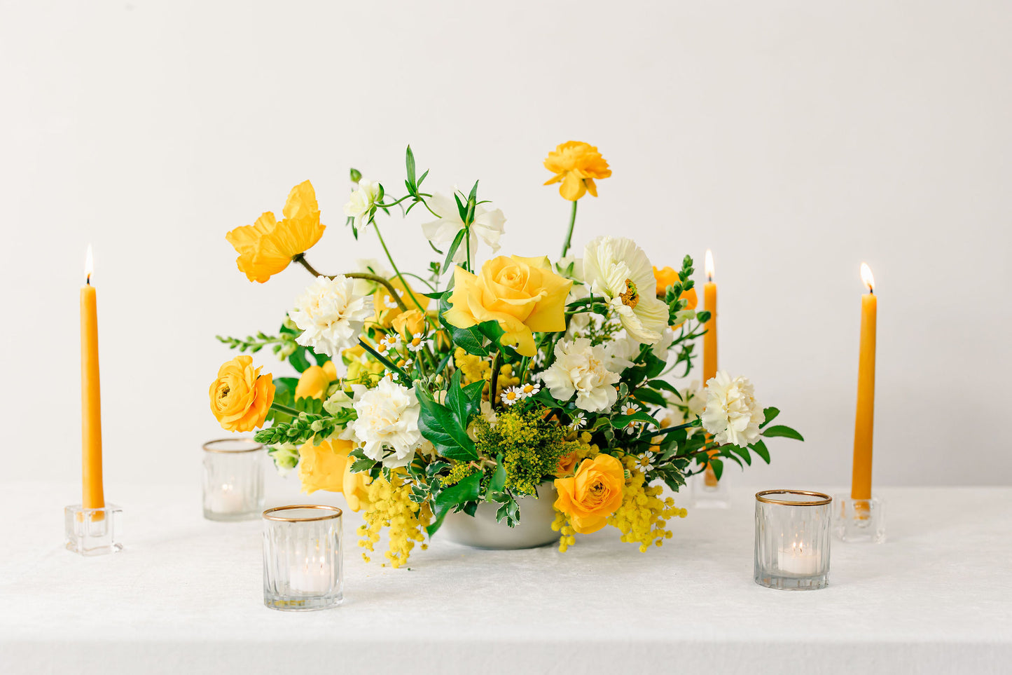 Floral arrangement with yellow and white flowers and candles on a white surface