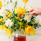 Honey being drizzled from a wooden dipper into a glass jar with a floral arrangement in the background.