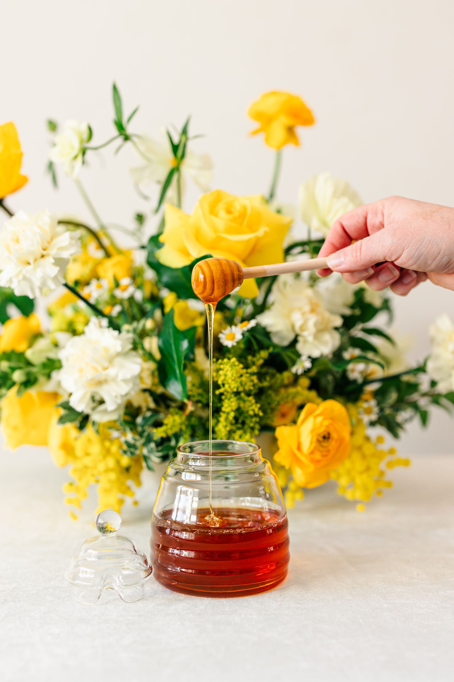Honey being drizzled from a wooden dipper into a glass jar with a floral arrangement in the background.