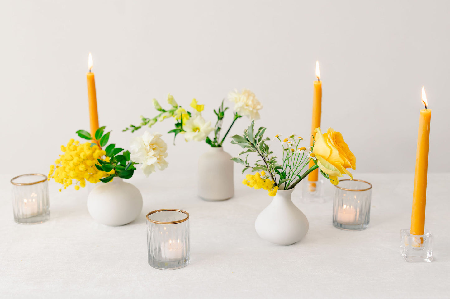 Yellow flowers in white bud vases with yellow candles on a white background