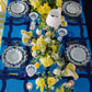 Decorative table setting with floral centerpieces, blue and white tablecloth, and patterned plates.