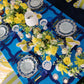Decorative table setting with blue and yellow tablecloth, floral arrangements, and white plates.