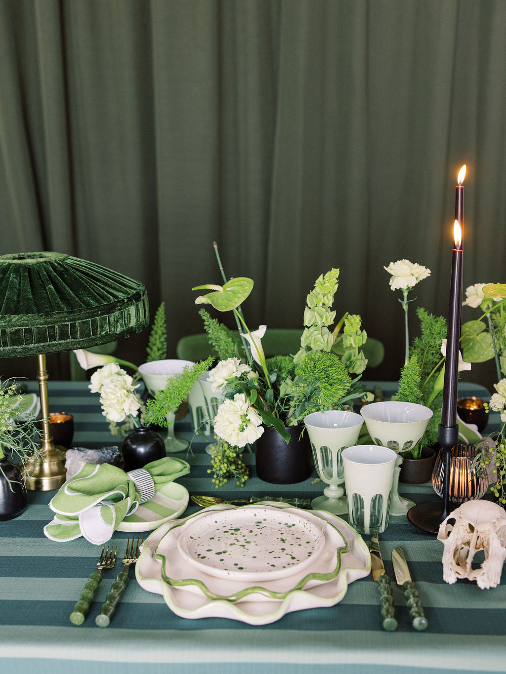 Elegant table setting with green and white decor, including plants and candles, against a dark curtain background.