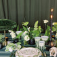 Elegant table setting with green and white decor, including plants and candles, against a dark curtain background.